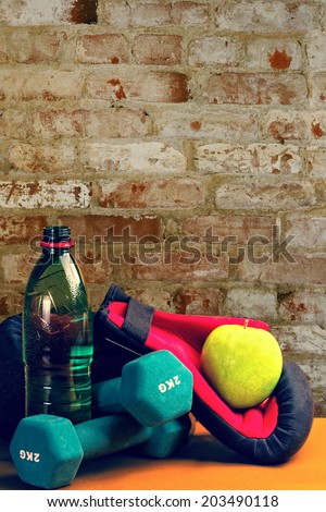 Composition presenting healthy lifestyle,with green apple,boxing gloves,bottle of water and dumbbells,against wall background.