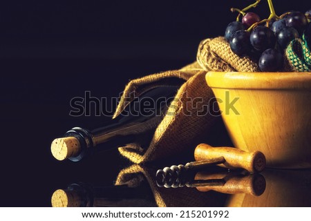 Close up of a wine bottle and dark grapes in wooden basket, against black background.
