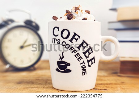 White coffee mug with cream, in front of old clock and books, on wooden table.