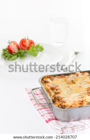 Lasagna in a baking dish in front of a plate with salad. A glass with water is present in the background