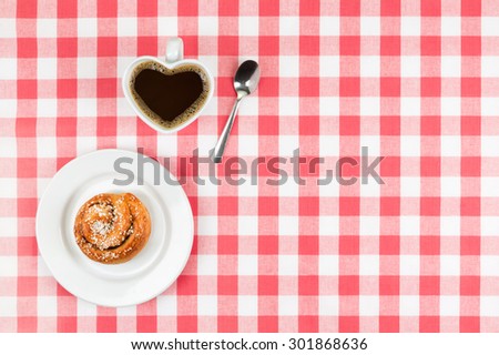 Heart shaped mug with coffee with a delicious cinnamon roll on red a checkered cloth with free space on the right side text etc according to your project.
