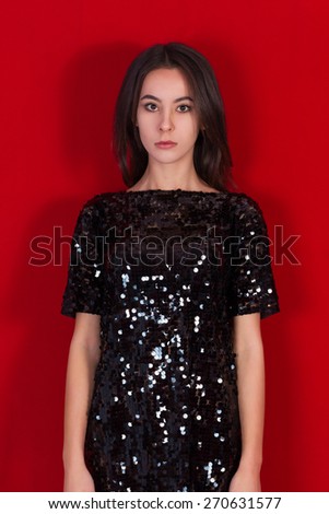 Beautiful brunette girl in a black dress.  Incredible young woman dressed in a little short shiny dress standing on a red background. Brunette with long black hair