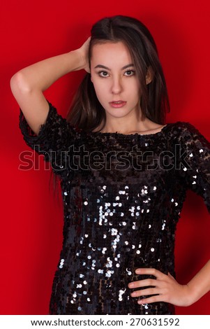 Beautiful brunette girl in a black dress.  Incredible young woman dressed in a little short shiny dress standing on a red background. Brunette with long black hair
