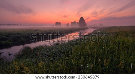 A small river at dawn in the mist among the meadows with beautiful clouds