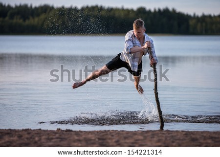 A young man is jumping in shallow water with the help of a wooden stick.