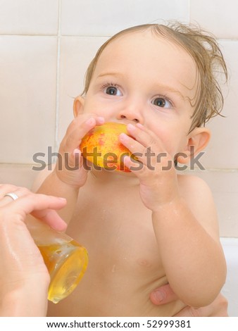 a one year old boy eats half of a nectarine in a bath tub; his mom is holding a glass of yellow juice