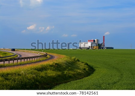 A road bend by green fields with a factory on the horizon