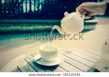 Hand poures green tea from white ceramic teapot into a matching tea cup, tinted image