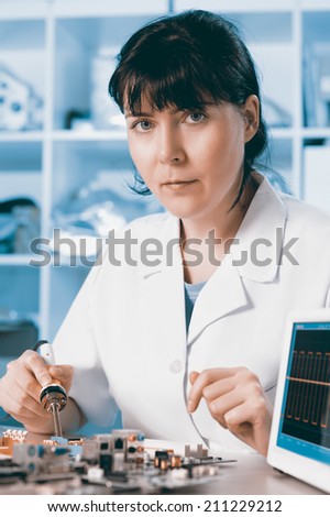 Young female tech soldering a circuit board looks at the viewer, toned image