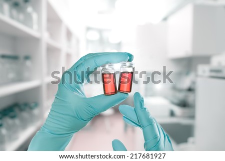 Two hands in nitril gloves hold red liquid samples in disposable plastic vials, laboratory interior out of focus