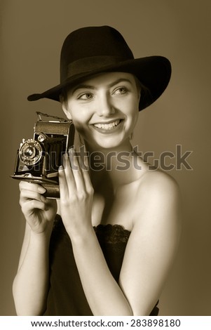 Attractive young lady in black hat with vintage portable camera in hands on neutral background. This image is toned for retro-look