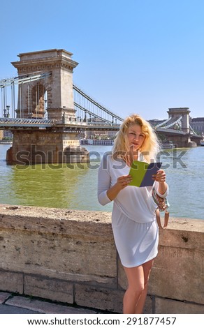 Blonde female tourist in white dress with book in hands poses in front of famous Chain Bridge in Budapest