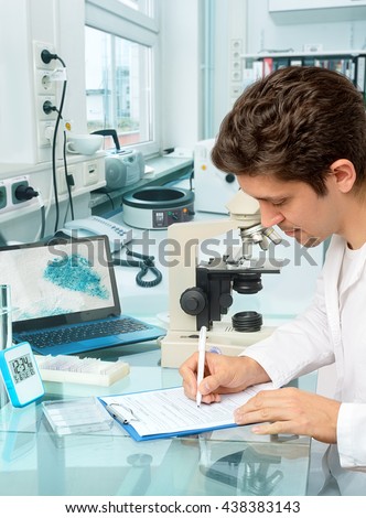 Young male scientist or tech makes notes in histopathology laboratory with light microscope and a laptop. Shallow DOF, focus on the right hand,