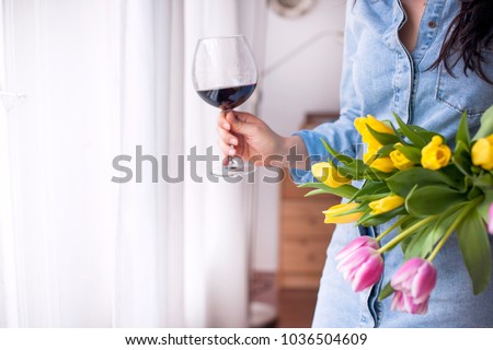 A woman with a glass of red wine, holding a bouquet of fresh tulips of yellow and pink in her hands. A gift for the beloved. Near the window. Postcard, place for text.