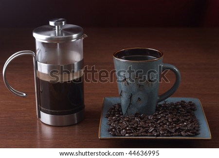 A coffee press sits on a table with a coffee cup and coffee beans