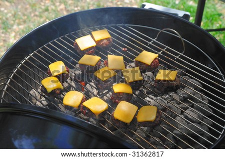 Several small hamburgers on the grill over hot charcoal covered with cheddar cheese