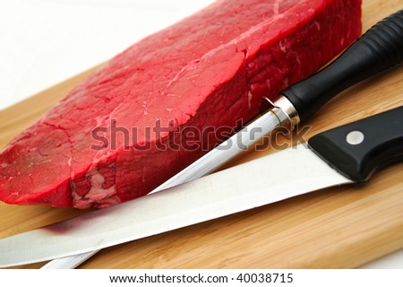 A large uncooked steak on a cutting board with butcher knife and sharpener on a white background