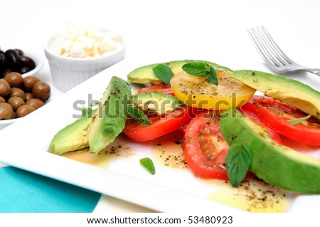 Red and yellow sliced tomatoes with  avocado, fresh oregano leaves with an olive oil and raspberry vinaigrette dressing on a square white plate with green and black olives in small bowls
