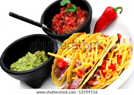 Three hard shell taco\'s with sides of fresh salsa and guacamole served on a white plate and a single red chili pepper on the side.