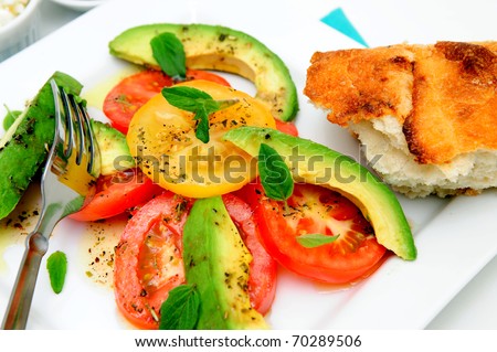 Fresh red and yellow tomatoes, sliced avocado, basil leaves and raspberry vinaigrette dressing on a square white plate and piece of artisan bread
