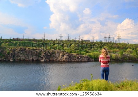 The girl stands near the river and looks at a power line
