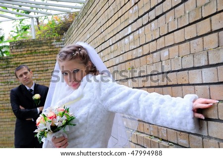 The groom looks at the bride. The bride leans on a brick wall and looks in the cam