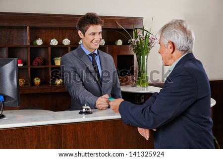 Smiling receptionist behind counter in hotel giving key card to senior guest