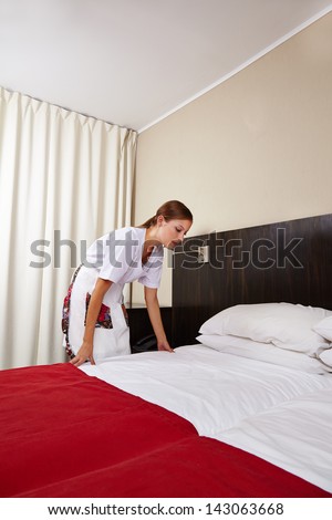 Young housekeeping maid cleaning bed in hotel room