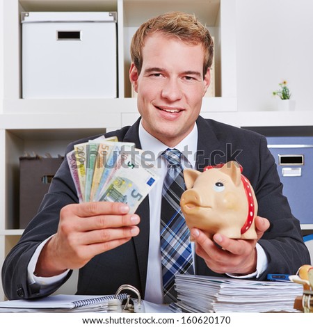 Happy business man in his office holding Euro money bills and a piggy bank