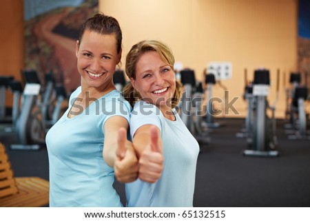 Two happy sporty women standing in a gym and holding their thumbs up