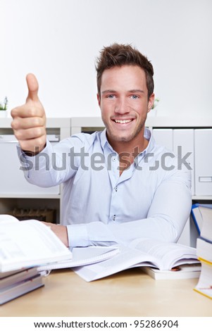 Successful university student holding thumbs up with books at desk