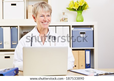 Elderly female doctor sitting smiling at laptop computer in her office