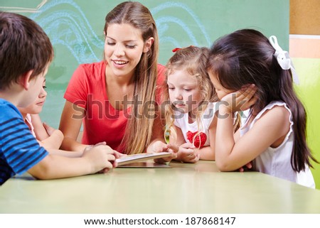 Many children at table reading a book with kindergarten teacher in preschool