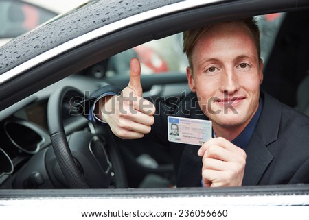 Young proud man in a car holding drivers licence and his thumps up