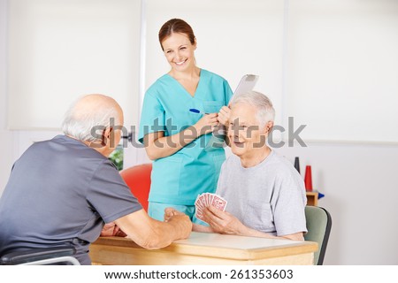 Two old men playing cards in a nursing home with smiling nurse watching