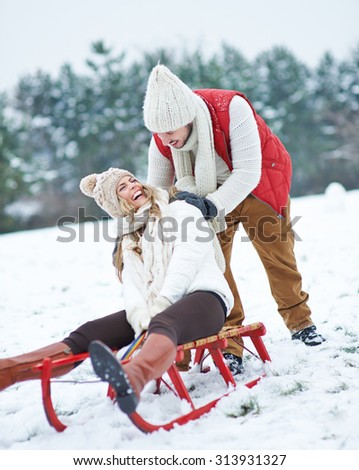 Happy man pushing laughing woman on sled through snow in winter