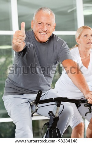 Happy senior man holding thumbs up on bike in fitness center