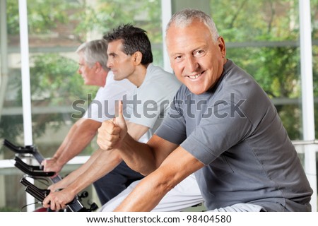 Happy senior man on bike holding thumbs up in fitness center