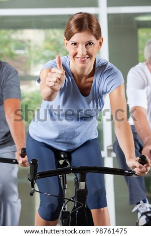 Elderly woman in fitness center on bike holding thumbs up