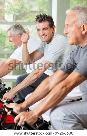 Happy senior man in gym on bike holding thumbs up