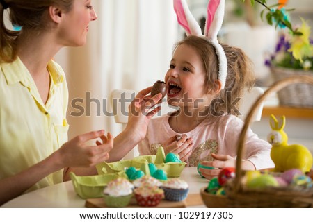 Mother and daughter celebrating Easter, eating chocolate eggs. Happy family holiday. Cute little girl in bunny ears laughing, smiling and having fun.