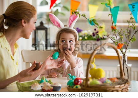 Mother and daughter celebrating Easter, eating chocolate eggs. Happy family holiday. Cute little girl with funny face in bunny ears laughing, smiling and having fun.