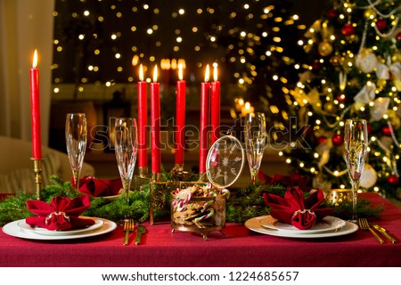 Beautiful served table with candles, Red tablecloth and napkins, white china, gold cutlery, crystal champagne glasses. Living room decorated with lights and Christmas tree. Holiday setting