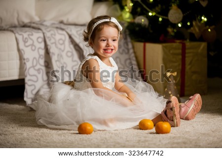 Cute happy little girl in beautiful white dress sitting on the carpet next to the christmas tree. Christmas tree with lights and gift boxes on the background. She is smiling