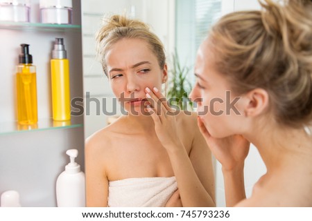 Young beautiful woman touching skin in bathroom. Unhappy girl standing in towel, looking in the mirror, checking dry irritated skin, puffiness and dark circles under eyes. Morning skincare routine.