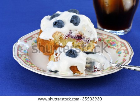 Blueberry and whipped cream topped cake on an antique floral plate.  A glass of ice coffee to the side.