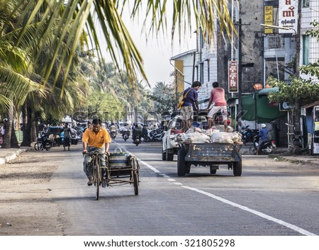 MYEIK, MYANMAR - FEBRUARY 20, 2015: A busy street at Myeik in the south of Myanmar. Two men are standing on the load area of a three-wheeler, a man is driving bycicle rikshaw.