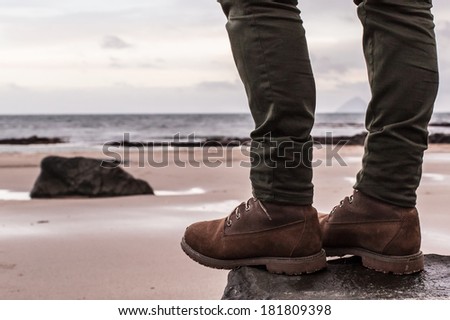 Women's 6-Inch Brown Waterproof Boots and skinny trousers on a rock with sand beach and ocean in the background. Scotland (UK)