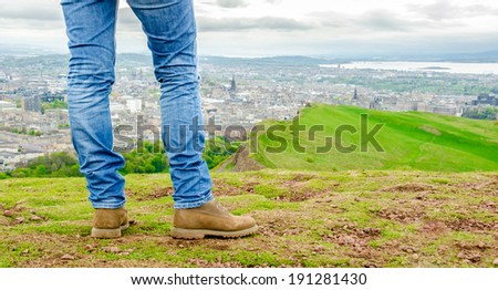 Focus on legs of a female model wearing skinny blue jeans and brown suede boots admiring Edinburgh landscape from the top of the Arthur\'s seat. Scotland, UK.