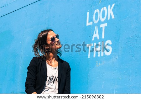 Hipster model wearing sunglasses posing next to a blue wall with the words \
